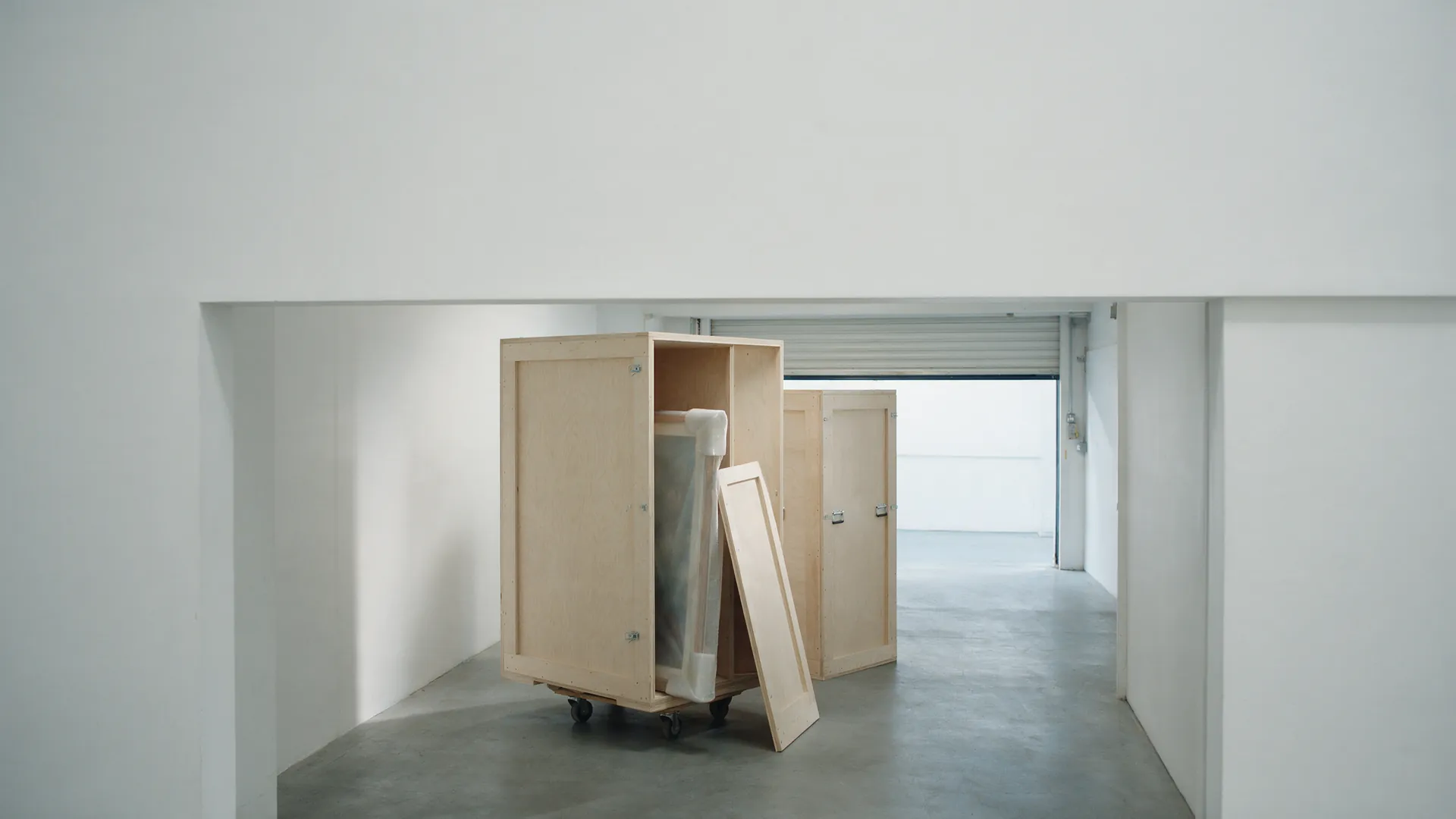 An open birch ply crate on a wheeled dolly in a fine art inspection bay, a wrapped work inside and the removed front panel leaning beside it, with nobody present