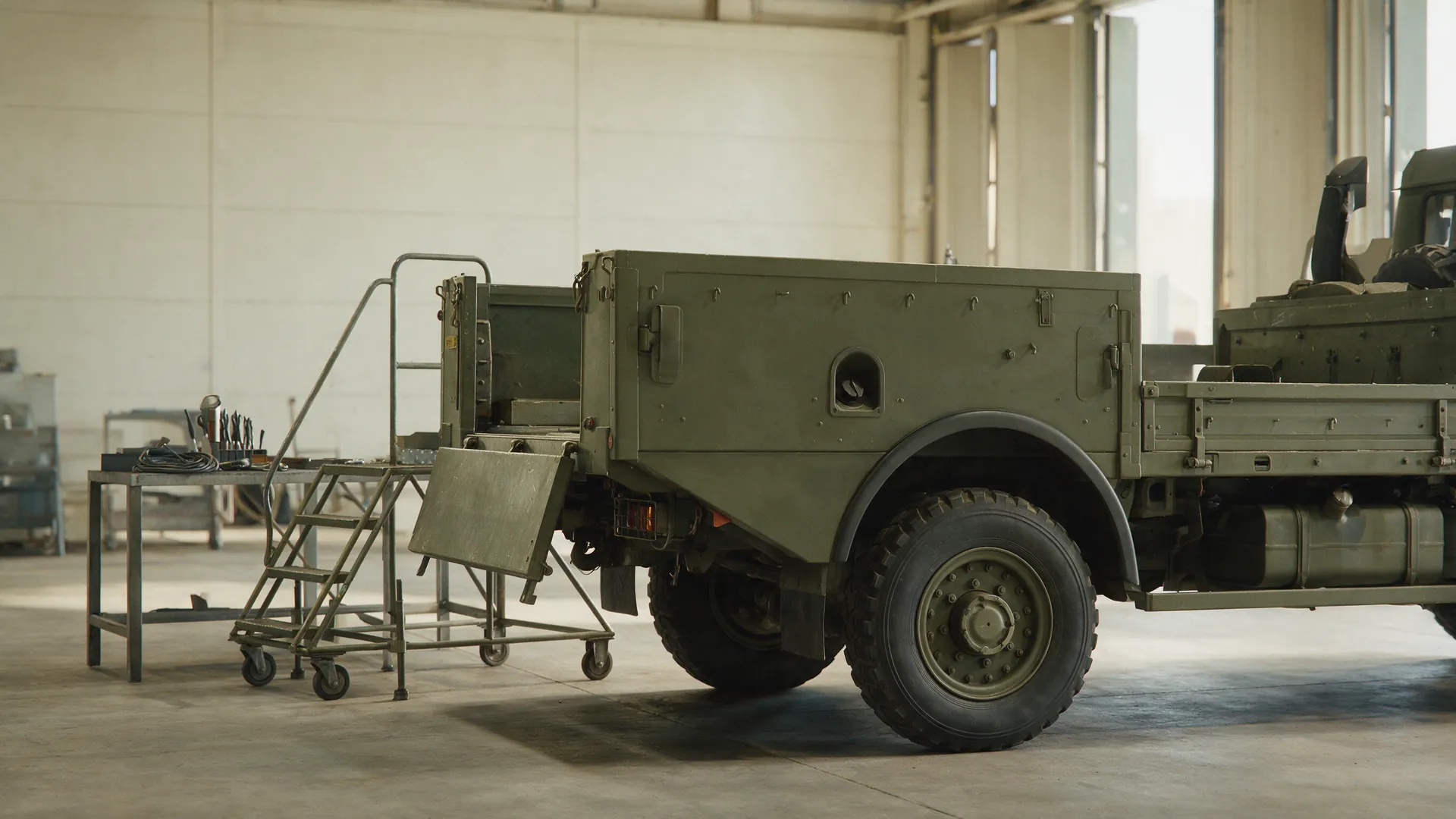 An olive support truck with its tailgate lowered in a maintenance hangar, an inspection platform and tool bench alongside, with nobody present